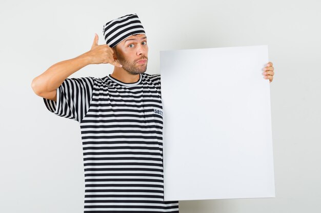Hombre joven que muestra el gesto del teléfono, sosteniendo el lienzo en blanco en camiseta rayada, sombrero.