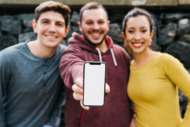 Hombre joven que muestra la pantalla vacía del teléfono inteligente mientras está de pie cerca de amigos multirraciales