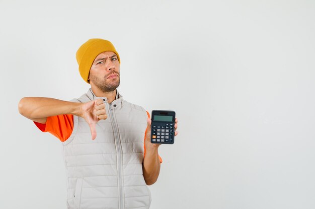 Hombre joven que muestra el pulgar hacia abajo mientras sostiene la calculadora en camiseta, chaqueta, sombrero y parece insatisfecho.