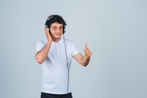 Hombre joven que muestra el pulgar hacia arriba mientras escucha música en una camiseta blanca y parece confiado. vista frontal.