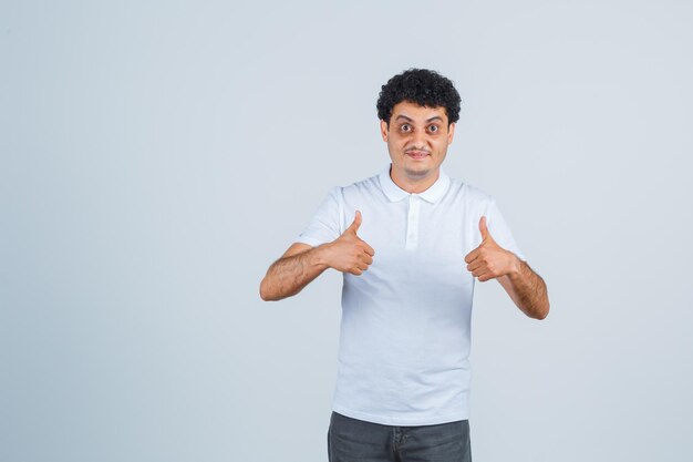 Hombre joven que muestra los pulgares para arriba con ambas manos en pantalones vaqueros y camiseta blanca y parece feliz. vista frontal.