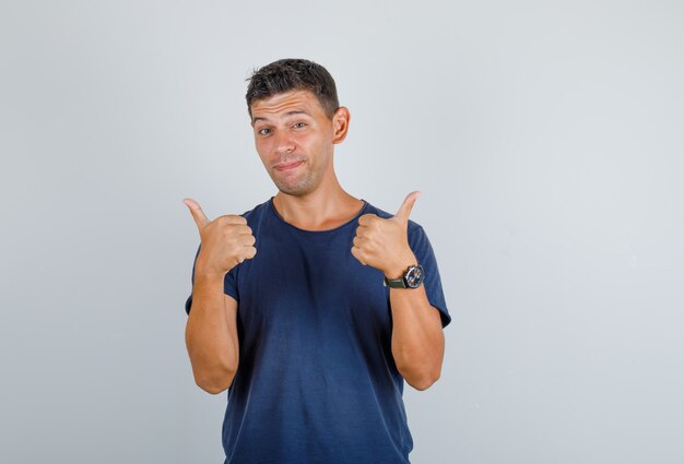 Hombre joven que muestra los pulgares para arriba en la camiseta azul oscuro y parece contento. vista frontal.