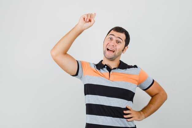 Hombre joven que muestra el signo de la nueva idea mientras pone la mano en la cintura en una camiseta y parece esperanzado. vista frontal.