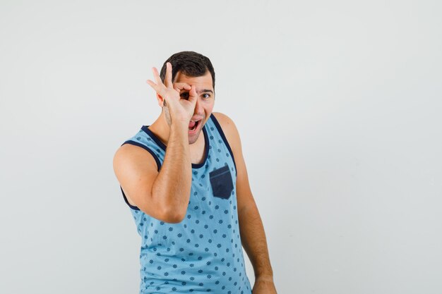 Hombre joven que muestra el signo ok en el ojo en camiseta azul y parece feliz. vista frontal.