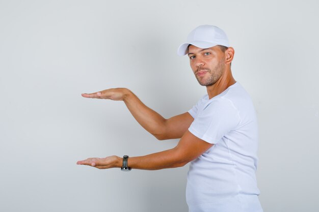 Hombre joven que muestra el signo de tamaño con las manos en camiseta blanca, gorra, vista frontal.