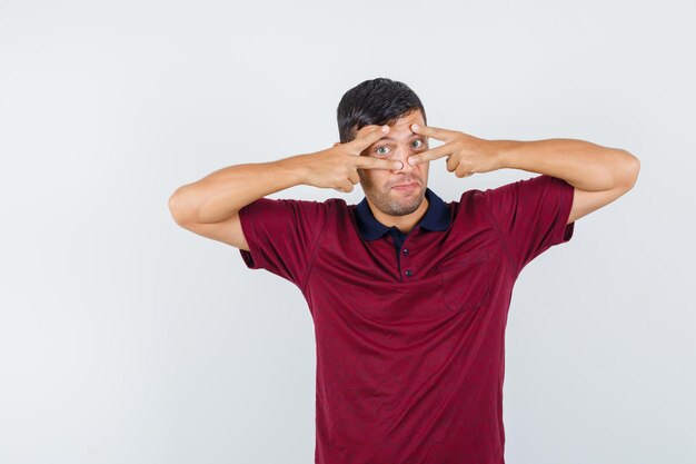 Hombre joven que muestra el signo v en los ojos en una camiseta y se ve lindo, vista frontal.