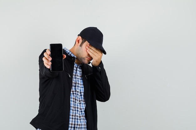 Hombre joven que muestra el teléfono móvil, sosteniendo la mano en los ojos en camisa, chaqueta, gorra, vista frontal.