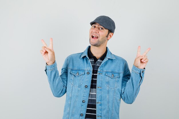 Hombre joven que muestra v-sign en chaqueta, gorra y mirando alegre, vista frontal.