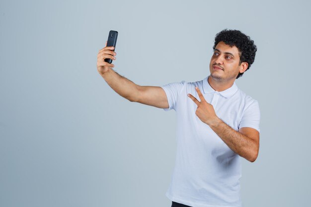 Hombre joven que muestra V-sign mientras toma selfie en camiseta blanca y luce orgulloso, vista frontal.