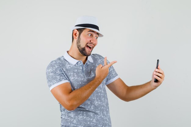 Hombre joven que muestra v-sign mientras toma selfie en camiseta, sombrero y mirando alegre, vista frontal.