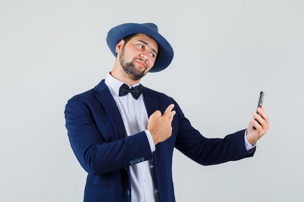 Hombre joven que muestra v-sign mientras toma selfie en traje, sombrero y luce optimista, vista frontal.