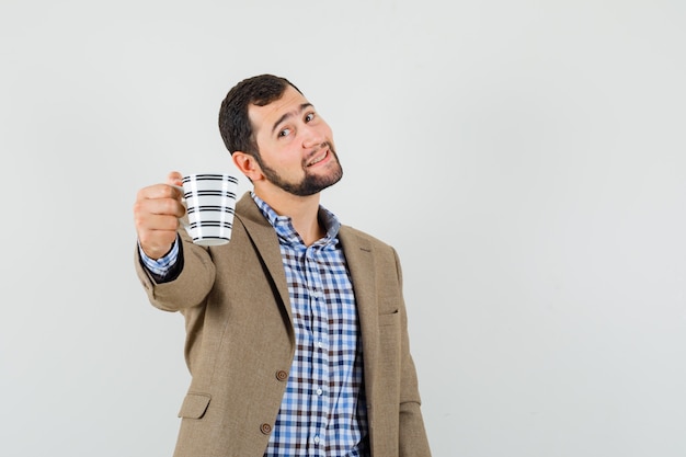 Hombre joven que ofrece una taza de café en camisa, chaqueta y mirada suave. vista frontal.