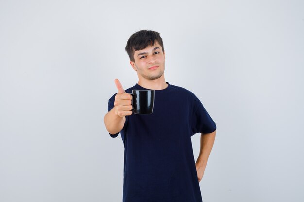 Hombre joven que ofrece una taza de café en camiseta negra y parece orgulloso. vista frontal.