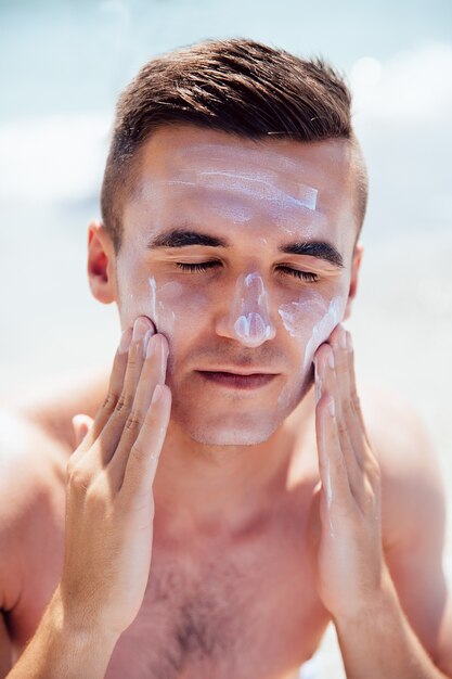 Hombre joven que pone la crema que broncea en su cara, tomando el sol en la playa. Cuidado de la salud.