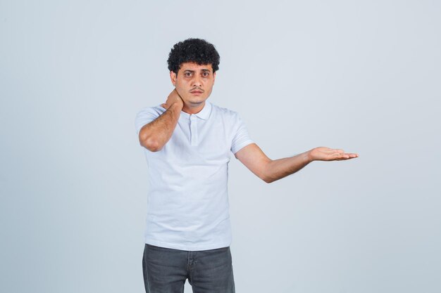 Hombre joven que pone la mano en el cuello, tiene dolor de cuello y estira la mano hacia la derecha en una camiseta blanca y pantalones vaqueros y parece agobiado. vista frontal.