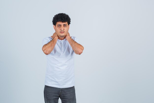 Hombre joven que pone las manos en el cuello, tiene dolor de cuello en camiseta blanca y jeans y parece acosado, vista frontal.