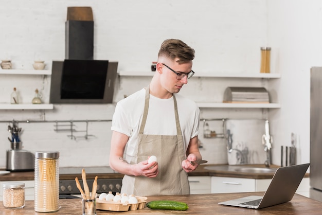 Hombre joven que prepara la comida mirando su computadora portátil en la mesa