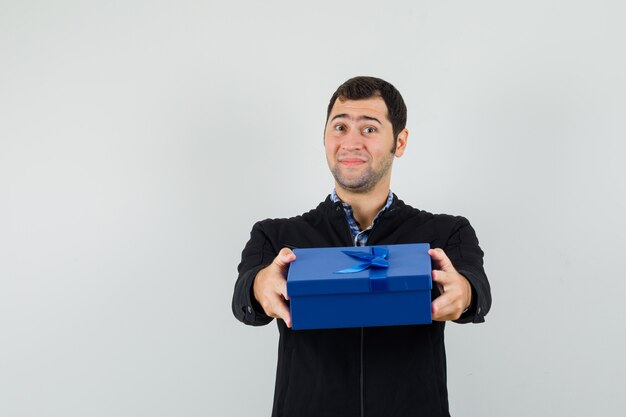 Hombre joven que presenta caja de regalo en camisa, chaqueta y mirando suave, vista frontal.