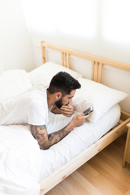 Hombre joven que se relaja en cama usando el teléfono móvil