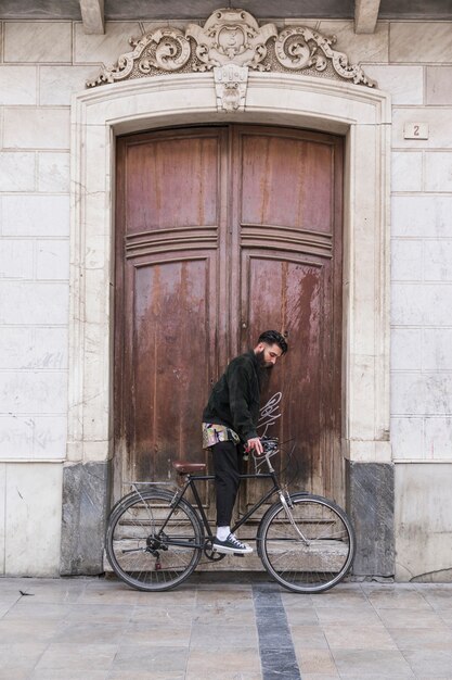 Hombre joven que se sienta en la bicicleta delante de la puerta de madera del vintage