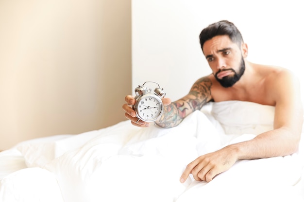 Hombre joven que se sienta en la cama que sostiene el despertador
