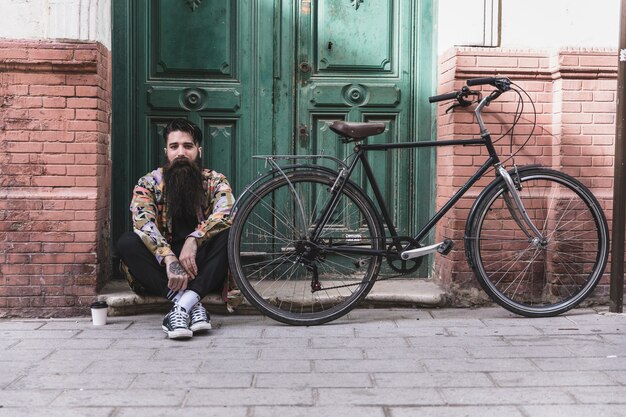 Hombre joven que se sienta cerca de la bicicleta con la taza de café para llevar delante de la pared verde de madera