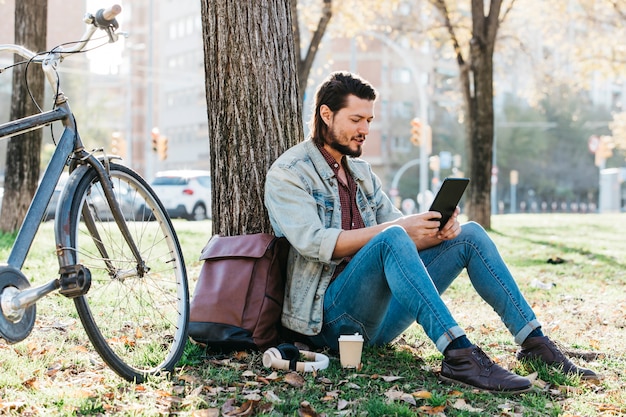Hombre joven que se sienta debajo del árbol usando el teléfono móvil en el parque con la taza de papel del café para llevar