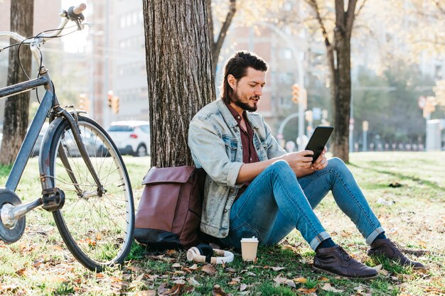 Hombre joven que se sienta debajo del árbol usando el teléfono móvil en el parque con la taza de papel del café para llevar