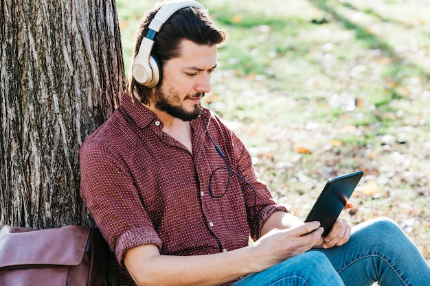 Hombre joven que se sienta debajo de la música que escucha del árbol en el auricular a través del teléfono móvil