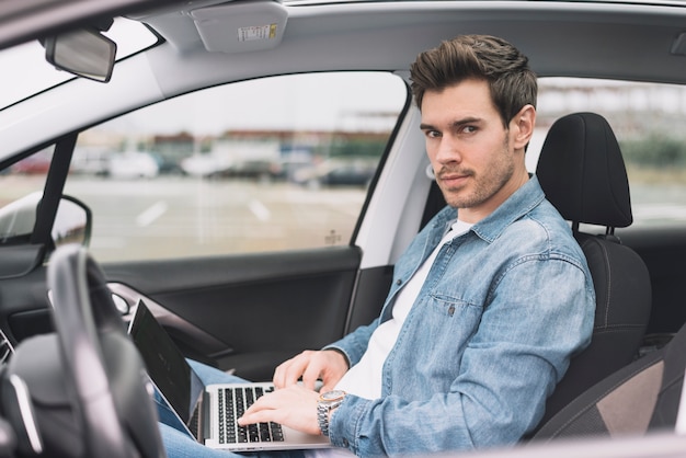 Hombre joven que se sienta dentro del coche moderno con la computadora portátil que mira la cámara