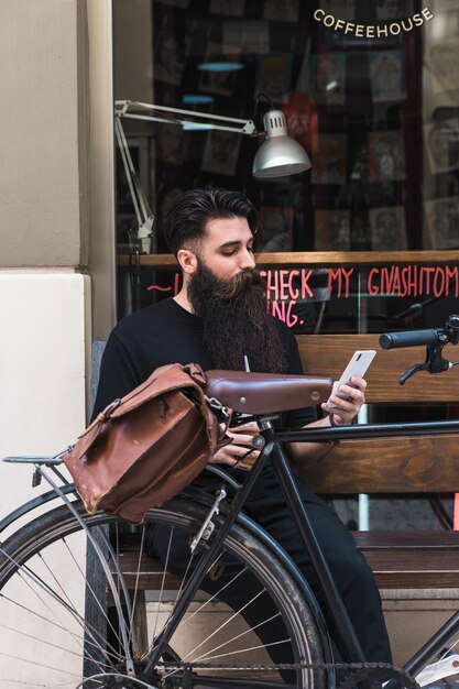 Hombre joven que se sienta fuera del café en banco con la bicicleta usando el teléfono móvil