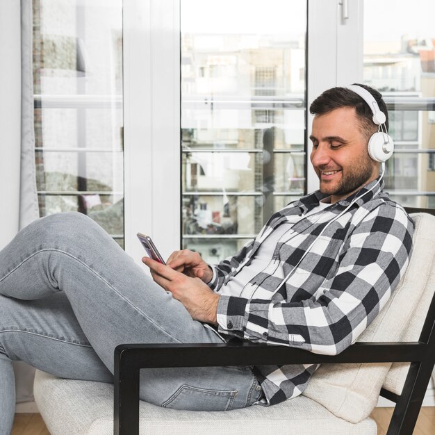 Hombre joven que se sienta en música que escucha de la silla en el auricular a través del teléfono móvil