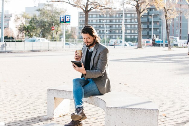 Hombre joven que se sienta en el parque de la ciudad que mira el teléfono móvil que sostiene la taza de café para llevar