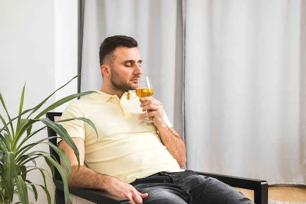 Hombre joven que se sienta en la silla que huele la bebida en copa de vino