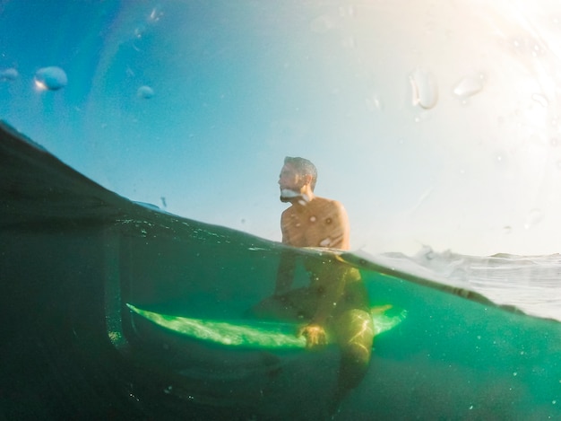 Hombre joven que se sienta en la tabla hawaiana en el mar azul