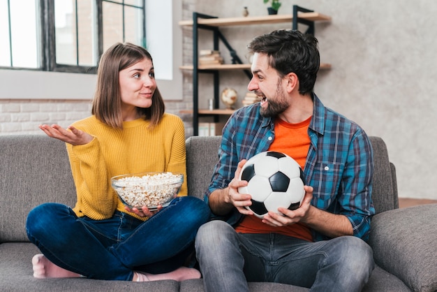 Hombre joven que sostiene el balón de fútbol en la mano que mira a su novia que sostiene el cuenco de palomitas de maíz