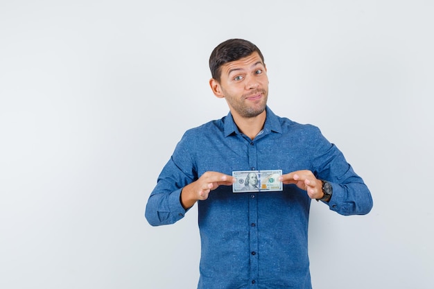 Hombre joven que sostiene el billete de un dólar en camisa azul y que parece afortunado. vista frontal.