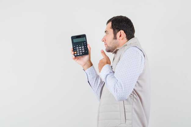 Hombre joven que sostiene la calculadora en una mano mientras muestra el pulgar hacia arriba en la chaqueta beige y la gorra y mira pensativo. vista frontal.