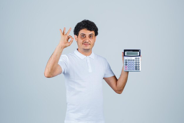 Hombre joven que sostiene la calculadora mientras muestra un gesto aceptable en camiseta blanca y parece confiado. vista frontal.