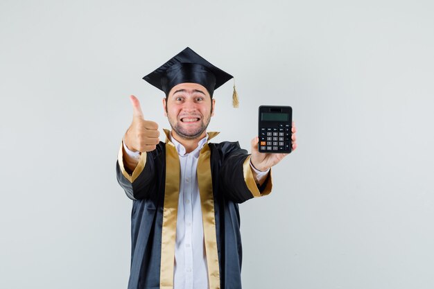 Hombre joven que sostiene la calculadora, mostrando el pulgar hacia arriba en uniforme de posgrado y mirando alegre, vista frontal.