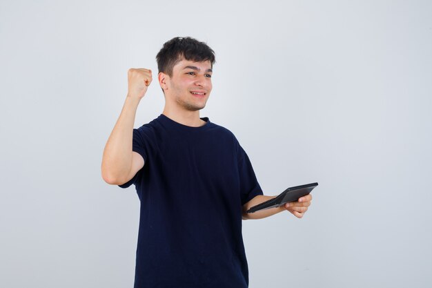 Hombre joven que sostiene la calculadora, que muestra el gesto del ganador en camiseta negra y parece afortunado. vista frontal.