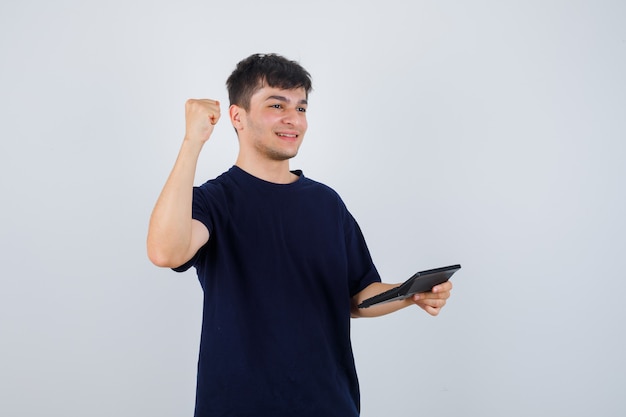 Hombre joven que sostiene la calculadora, que muestra el gesto del ganador en camiseta negra y parece afortunado. vista frontal.