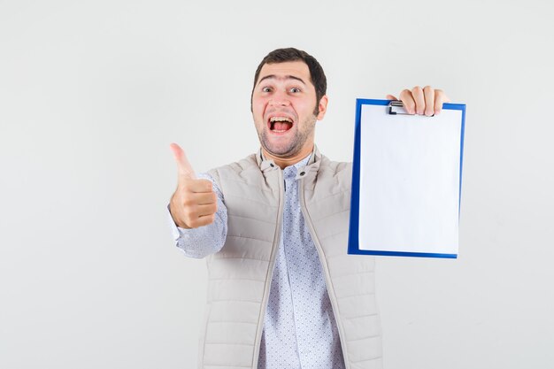 Hombre joven que sostiene el cuaderno y que muestra el pulgar hacia arriba en la chaqueta beige y que parece optimista. vista frontal.