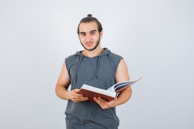 Hombre joven que sostiene el libro mientras mira a la cámara en una sudadera con capucha sin mangas y se ve guapo. vista frontal.