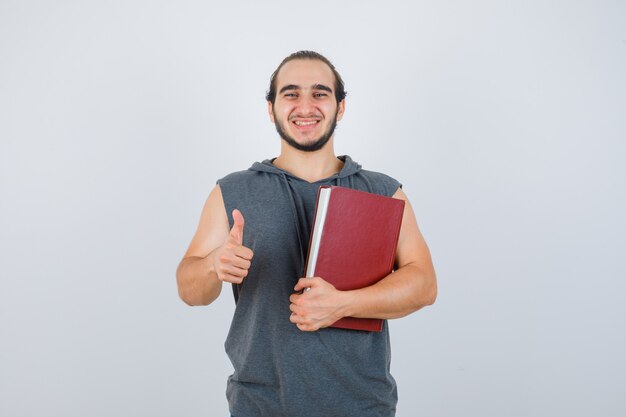 Hombre joven que sostiene el libro mientras muestra el pulgar hacia arriba en una sudadera con capucha sin mangas y parece feliz, vista frontal.