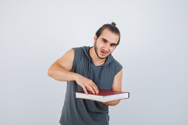 Hombre joven que sostiene el libro en una sudadera con capucha sin mangas y que mira confiado, vista frontal.