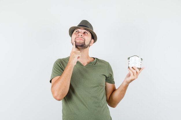 Hombre joven que sostiene el modelo de la casa mientras mira hacia arriba en camiseta verde, sombrero y aspecto soñador