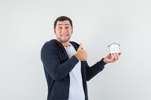 Foto gratuita hombre joven que sostiene el modelo de la casa y que muestra el pulgar hacia arriba en una camiseta blanca y una sudadera con capucha negra con cremallera frontal y un aspecto optimista. vista frontal.