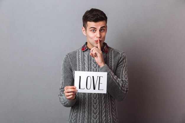 Hombre joven que se sostiene de papel con el texto del amor que muestra gesto del silencio.