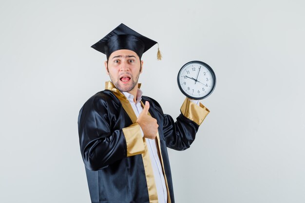 Hombre joven que sostiene el reloj de pared, mostrando el pulgar hacia arriba en la vista frontal uniforme graduado.